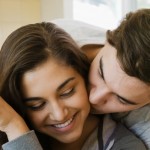 A smiling woman holds a mug while a man embraces her from behind, nuzzling her cheek.