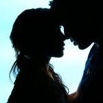 Silhouetted profiles of two people facing each other, noses nearly touching, against a bright, light blue background.