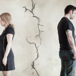 Woman and man stand apart, each holding half of a red broken heart, separated by a large crack in the wall behind them.