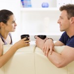 A woman and a man sit on a cream-colored couch, facing each other in profile, each holding a black mug.