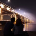At night, two people sit facing a brightly lit bridge over water, their backs to the viewer.