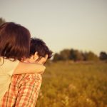 A woman hugs a man from behind in a field.