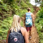 Two people hike a dirt trail through lush green foliage. The woman in the foreground has blonde hair in a ponytail and wears a backpack. A man in a tank top walks ahead of her.