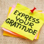 A stack of colorful sticky notes, with "Express Your Gratitude" written in black marker on the top yellow note, secured by a red push pin, rests on a textured burlap surface.