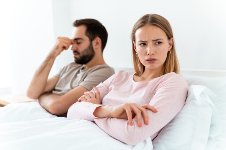 In bed, a woman with crossed arms and a furrowed brow sits beside a man who is touching his forehead.