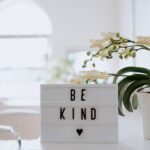 A white desk features a light box displaying "BE KIND" with a heart symbol, accompanied by a potted orchid.