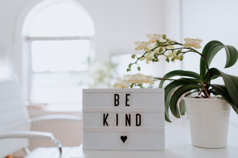 A white desk features a light box displaying "BE KIND" with a heart symbol, accompanied by a potted orchid.