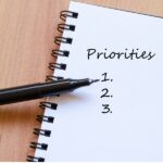 Spiral notebook with "Priorities" written on the top page, followed by numbered list 1, 2, 3, and a black pen pointing to the number 1.