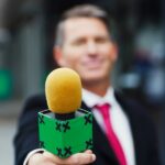 A man in a suit holds out a green microphone with a yellow windscreen.