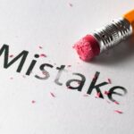 An orange pencil eraser rubs out the word "Mistake" on white paper, leaving pink eraser shavings.