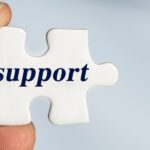 Fingers hold a white puzzle piece with the word "support" printed in dark blue.