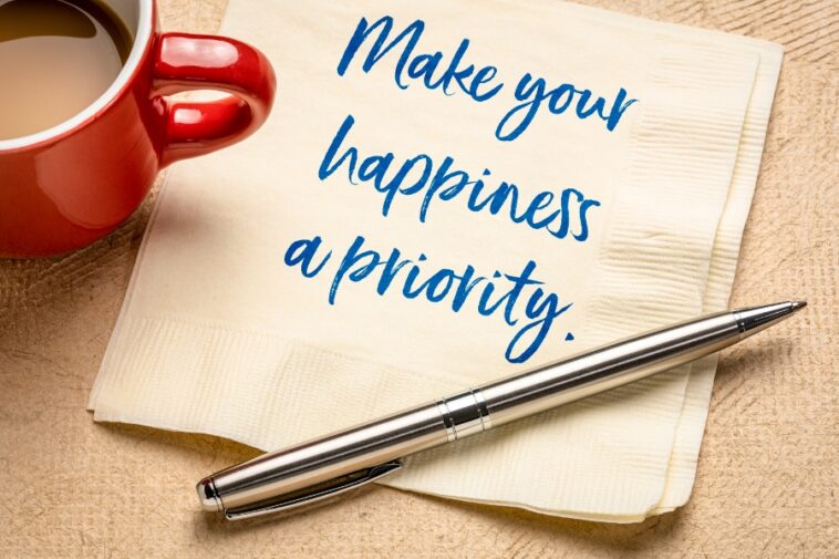 A red mug of coffee sits beside a napkin with the phrase "Make your happiness a priority" written in blue. A silver pen rests on the napkin.
