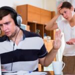 Man wearing headphones at a laptop raises his hand in a 'stop' gesture to a woman standing behind him with a concerned expression.