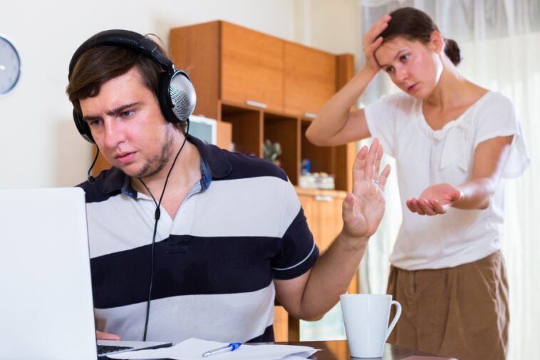 Man wearing headphones at a laptop raises his hand in a 'stop' gesture to a woman standing behind him with a concerned expression.