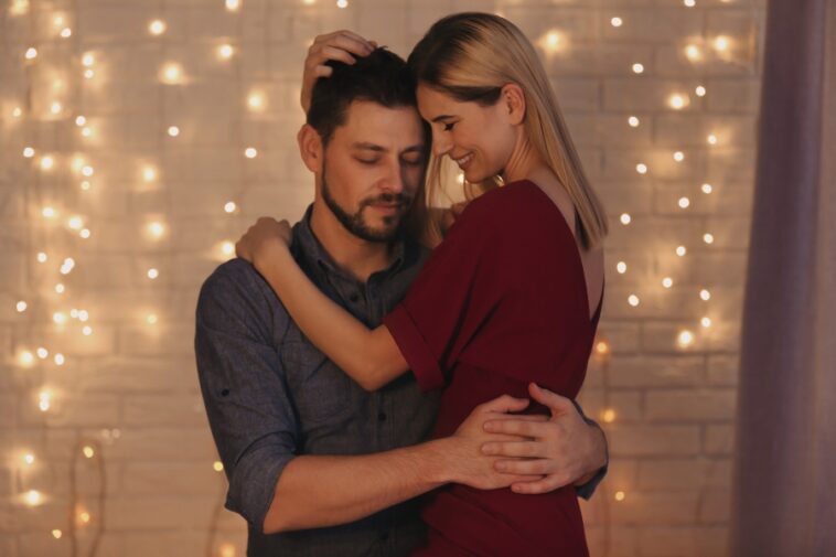 A woman in a red dress embraces a man in a blue shirt, standing in front of a wall with string lights.
