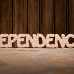 Wooden letters spelling "DEPENDENCY" sit on a textured surface in front of a wood-paneled wall.