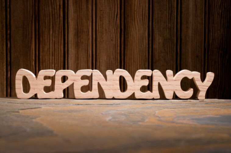 Wooden letters spelling "DEPENDENCY" sit on a textured surface in front of a wood-paneled wall.
