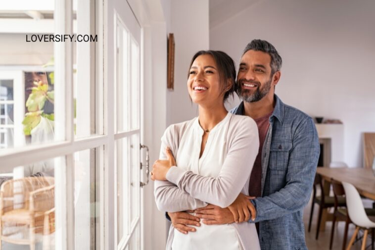 A smiling couple embraces while looking out a window.