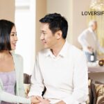 In a bright, airy living room, a young Asian couple sits on a sofa, holding hands and looking at each other. In the background, two older adults stand near a counter with a bottle and fruit.