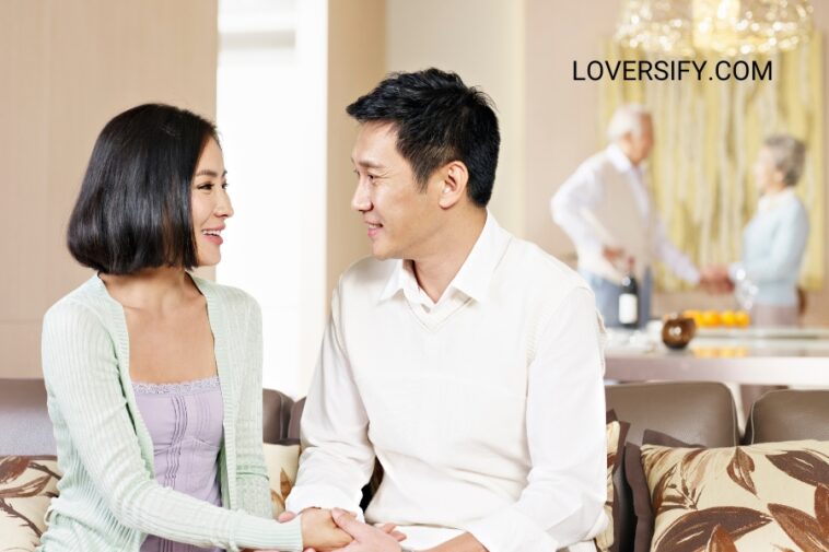 In a bright, airy living room, a young Asian couple sits on a sofa, holding hands and looking at each other. In the background, two older adults stand near a counter with a bottle and fruit.