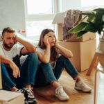 A man and woman sit on the floor, surrounded by moving boxes in a new home.