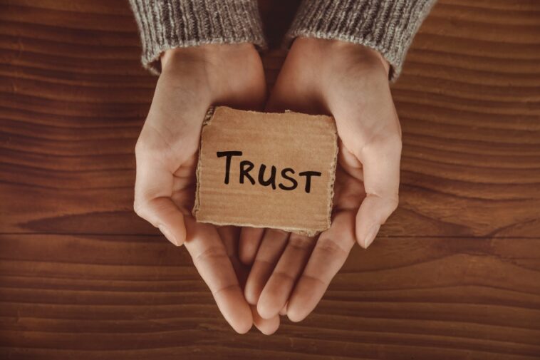 Hands wearing a sweater hold a cardboard square with the word "TRUST" written on it, over a wooden surface.