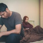 In a bedroom, a man sits on the edge of a bed with his head down, while a woman lies behind him with her arms crossed.