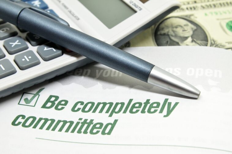 A pen rests on a form with a checked box next to the phrase "Be completely committed," a calculator and dollar bill are in the background.