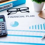 Overhead shot of a desk with a financial plan, calculator, glasses, pen, laptop, budget binder, and small potted plant.