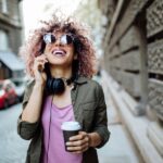 A smiling woman with curly pink hair, wearing sunglasses and headphones, talks on her phone while holding a coffee cup on a city street.