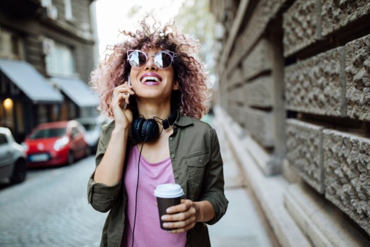 A smiling woman with curly pink hair, wearing sunglasses and headphones, talks on her phone while holding a coffee cup on a city street.