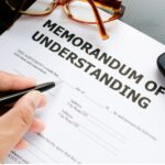 A hand holds a pen over a Memorandum of Understanding document, with glasses and a phone nearby.