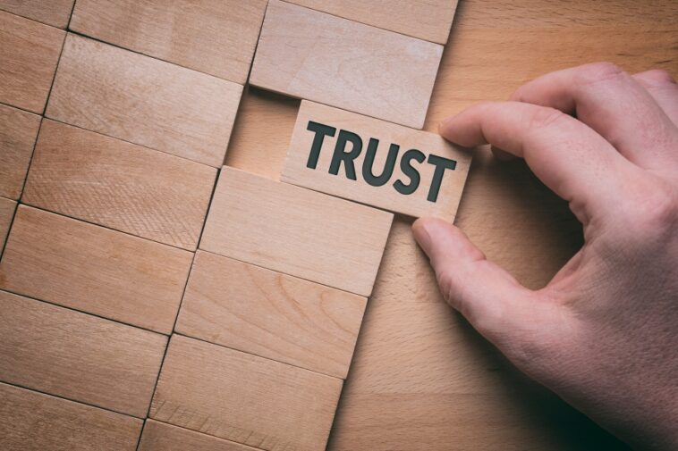 Hand placing a wooden block labeled "TRUST" into a puzzle of similar blocks.
