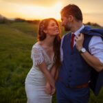 A bride and groom stand holding hands in a grassy field at sunset, gazing at each other.