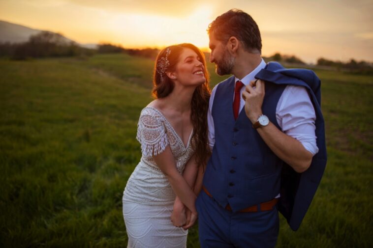 A bride and groom stand holding hands in a grassy field at sunset, gazing at each other.