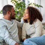 A smiling couple sits close together on a sofa, gazing at each other.