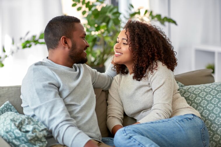 A smiling couple sits close together on a sofa, gazing at each other.