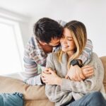 A man in a striped shirt hugs a smiling woman wearing a gray sweater on a couch.