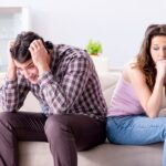 A man and woman sit on a sofa, backs touching, both looking downcast. The man has his hands on his head.