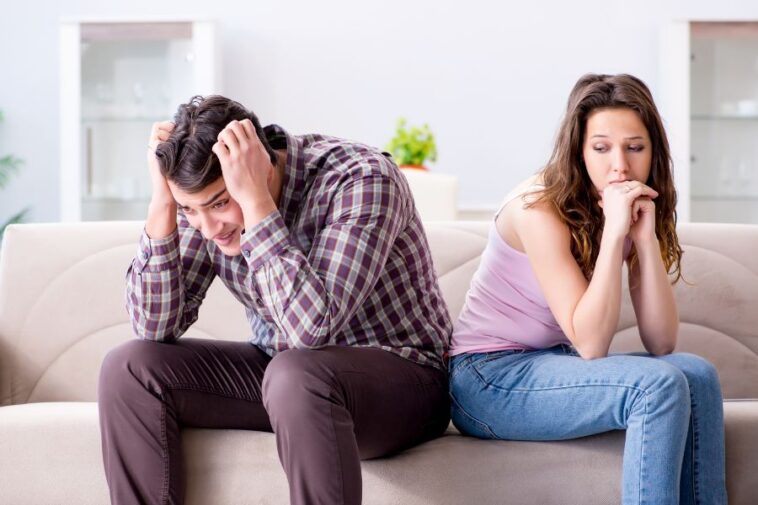 A man and woman sit on a sofa, backs touching, both looking downcast. The man has his hands on his head.