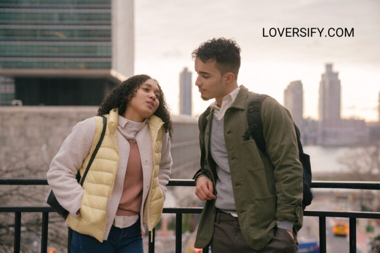 A young woman and man lean against a railing with a city skyline in the background.