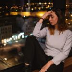 A woman sits by a large window at night, with city lights visible outside. She rests her head in her hand.