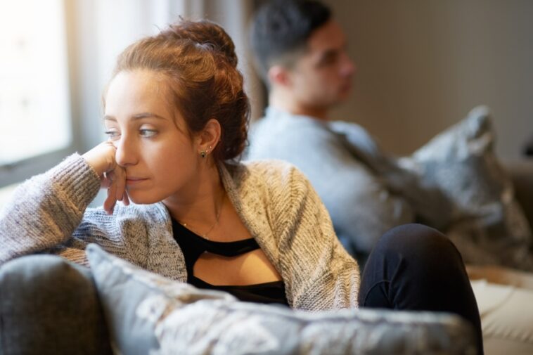 A woman sits on a couch, looking off to the side with her hand on her face, while a man sits blurred in the background.