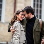 A smiling man with glasses embraces a smiling woman in a trench coat outdoors.