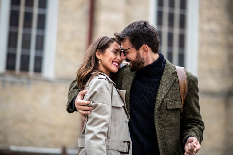 A smiling man with glasses embraces a smiling woman in a trench coat outdoors.
