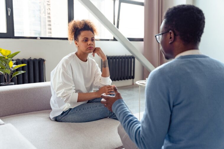 In a bright, modern room, a woman sits on a sofa, listening intently to a man facing her, gesturing with his hands.