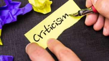 Hand holds a fountain pen over a yellow sticky note with the word "Criticism" written on it, with crumpled yellow and purple paper in the background.
