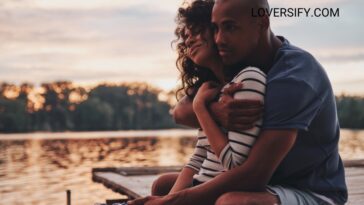 A man embraces a woman from behind while sitting on a dock with a lake and trees in the background at sunset.