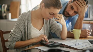 A woman uses a calculator while sitting at a table covered with papers, as a man stands behind her with his hand on his head.