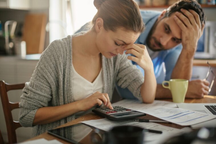 A woman uses a calculator while sitting at a table covered with papers, as a man stands behind her with his hand on his head.